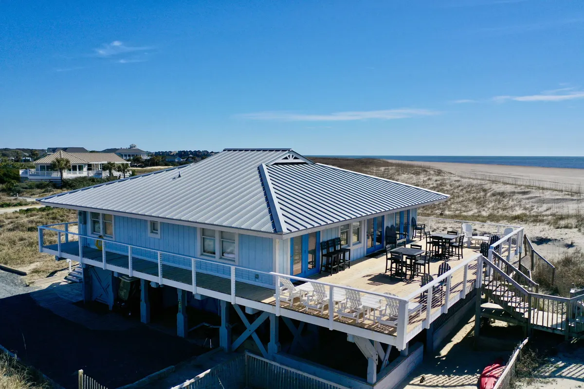 A wide view of the beach house showing its expansive deck directly overlooking the ocean and sandy beach. The deck is furnished for outdoor lounging and socializing, with the shoreline just steps away. The image conveys a relaxed, communal atmosphere perfect for group gatherings.