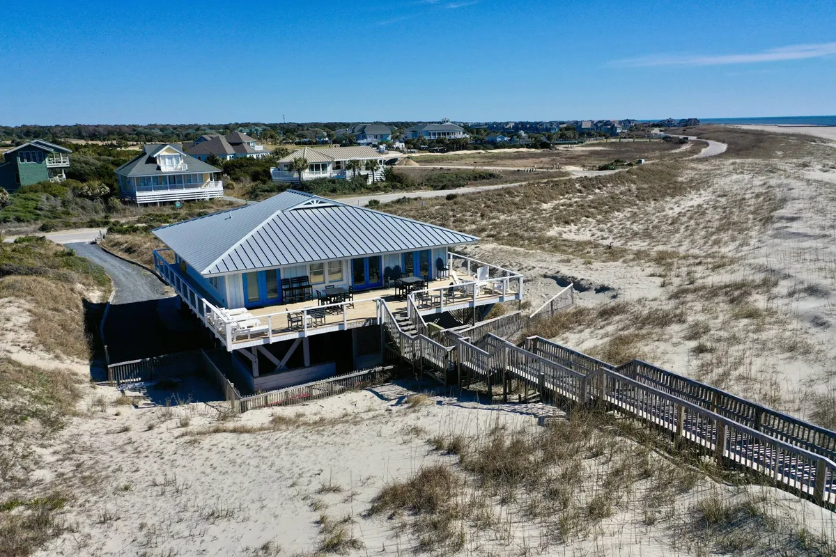 A charming beach house with a spacious deck overlooks the surrounding landscape, with a clear pathway leading directly to the beach. The deck area invites relaxation, socializing, and basking in coastal breezes, while the landscape emphasizes the natural, pet-friendly environment.