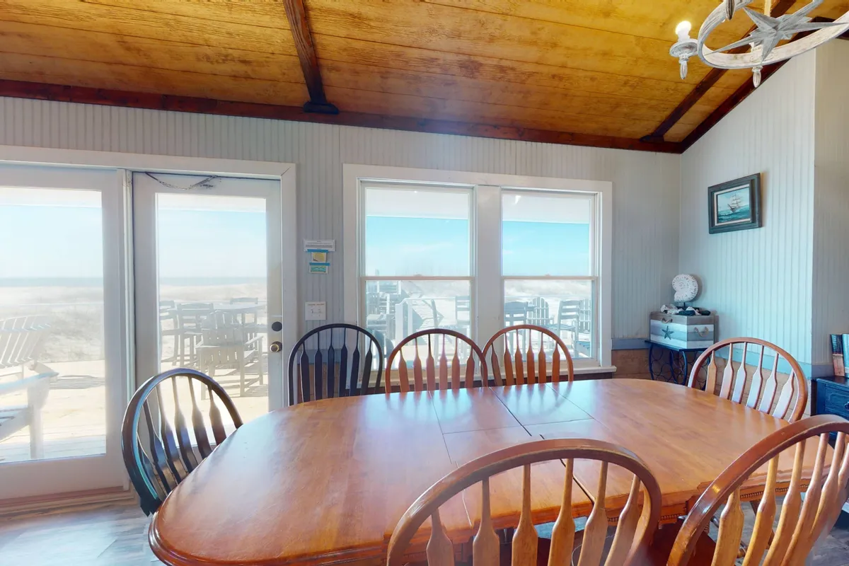 A large wooden dining table set near expansive windows, showcasing panoramic outdoor views. The room is warmly lit, with a chandelier overhead, and set up for group meals or game nights, mirroring the communal dining experiences described.
