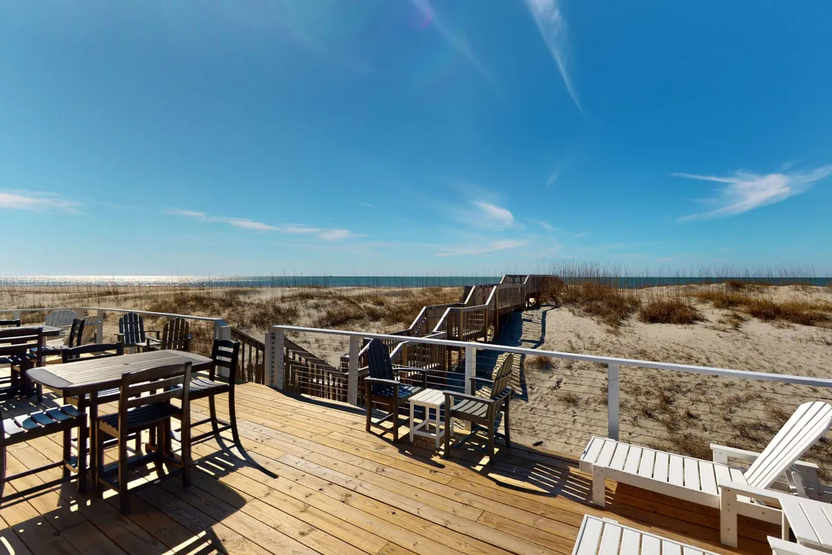 This image features a beachfront deck with comfortable seating overlooking a sweeping ocean view. A wooden walkway leads from the deck down toward the sandy beach, seamlessly connecting the outdoor living area to the shoreline. The relaxed, sunlit setting is perfect for gatherings, dining, or enjoying the tranquil sounds of the waves.