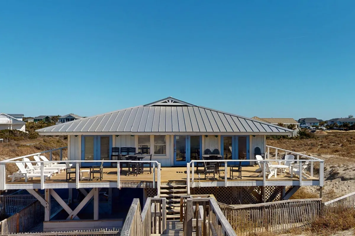 A broad, sun-drenched deck extends from the home, furnished with several seating arrangements and surrounded by sandy terrain. The unique roofline creates shaded spots, while the open design emphasizes the spaciousness and connection to the beach environment. The mood is relaxed, inviting visitors to linger outdoors from morning coffee to evening stargazing.