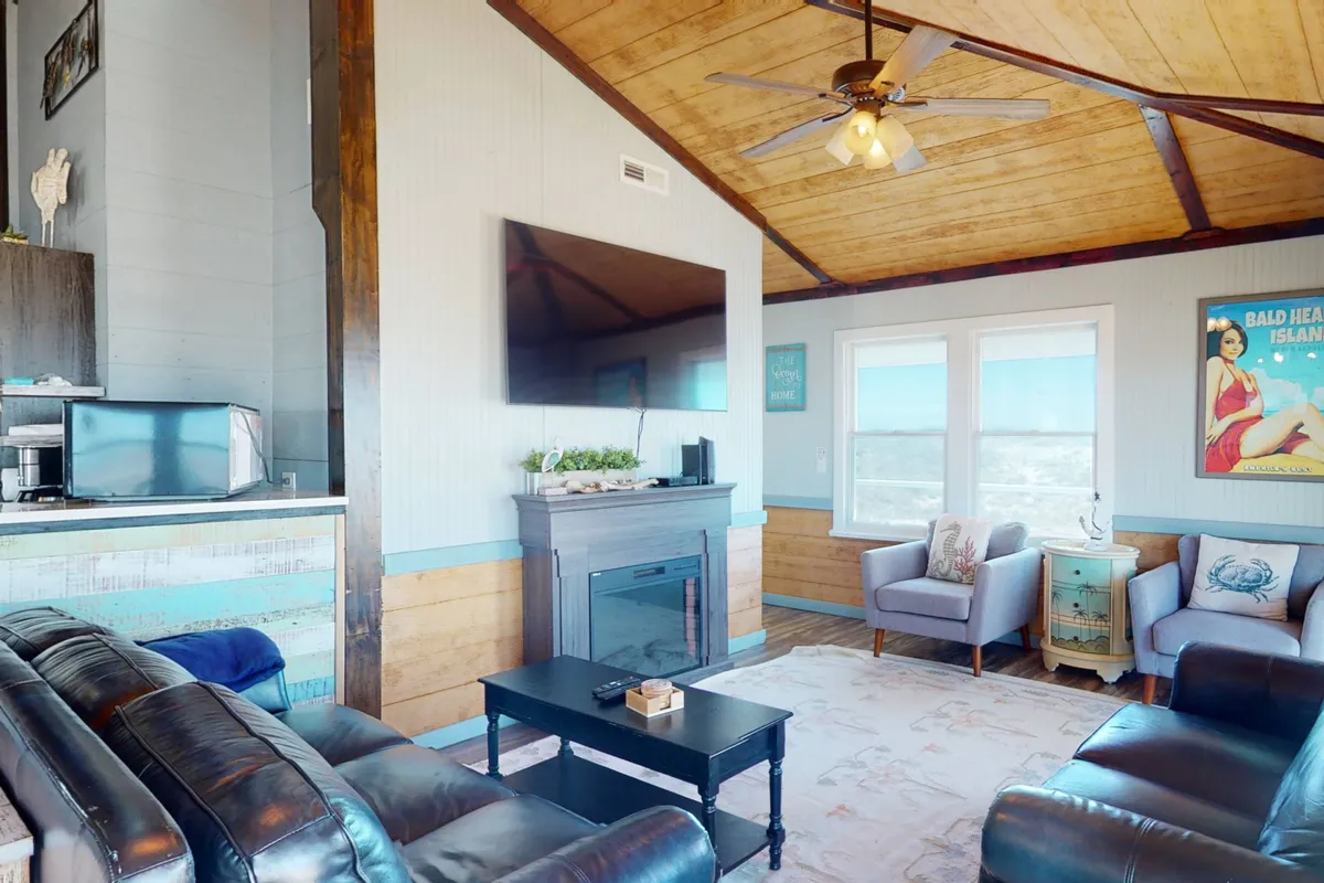 A spacious living room with a leather sofa (which doubles as a queen sleeper), a fireplace, large TV, decorative artwork, and windows with scenic views.