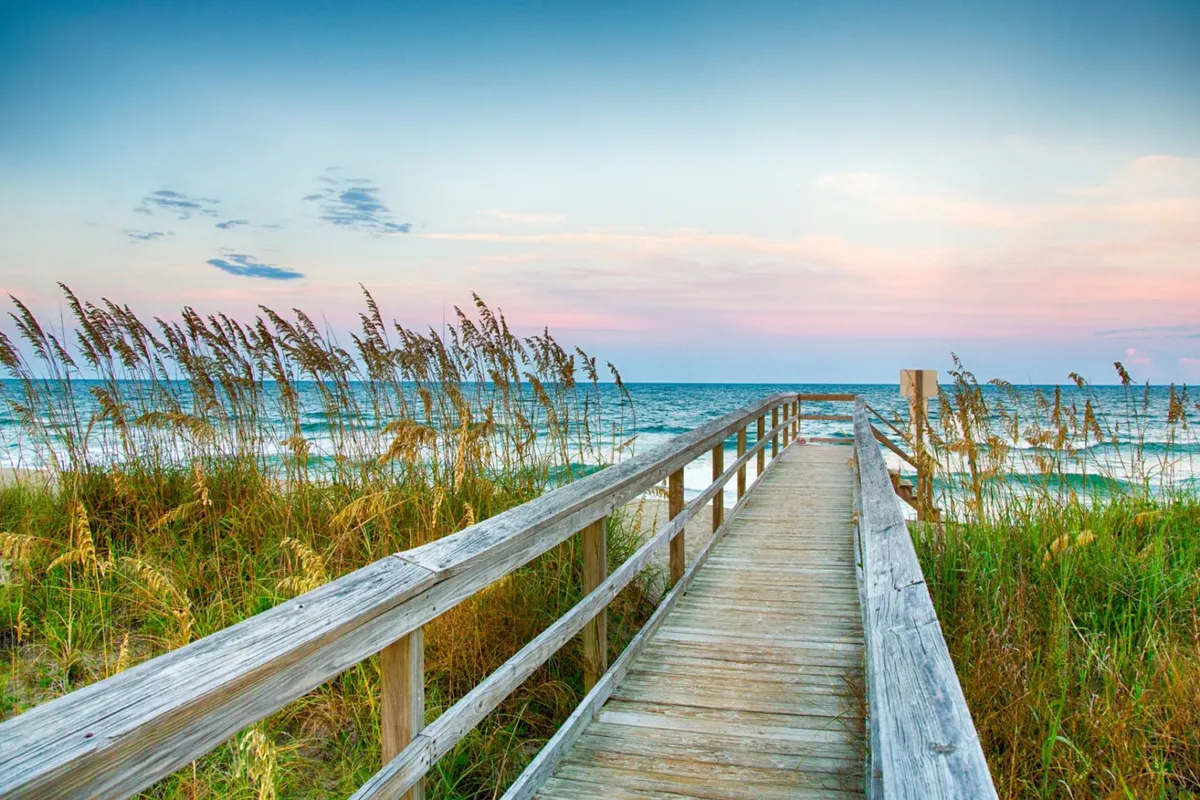 A long boardwalk leads across sand dunes to meet the ocean, with golden sunset hues bathing the scene. Dune grass and open water frame the path, evoking tranquility and opportunities for serene reflection and wildlife observation.
