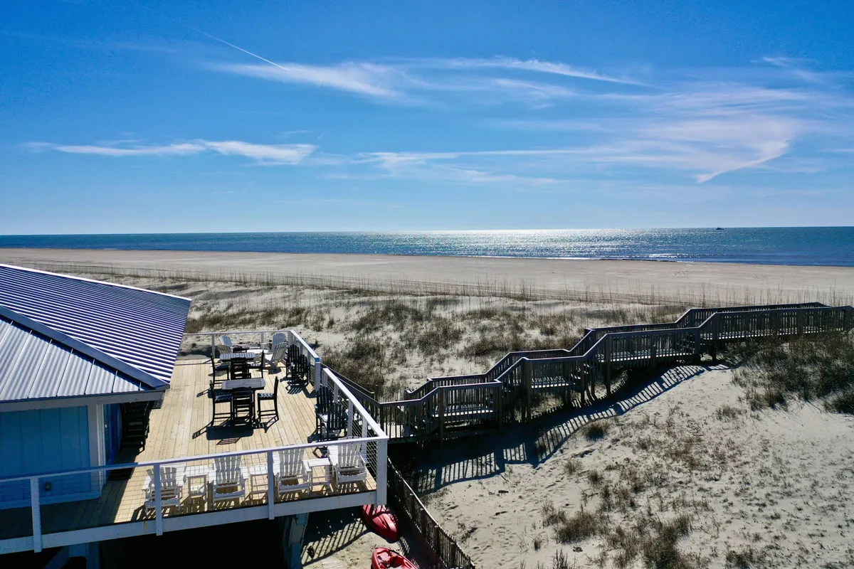 A serene view of a beachfront deck area with steps leading directly to the sand. The scene includes space for outdoor dining and activities, reinforcing a seamless blend of indoor-outdoor family fun and relaxation just steps from the ocean.