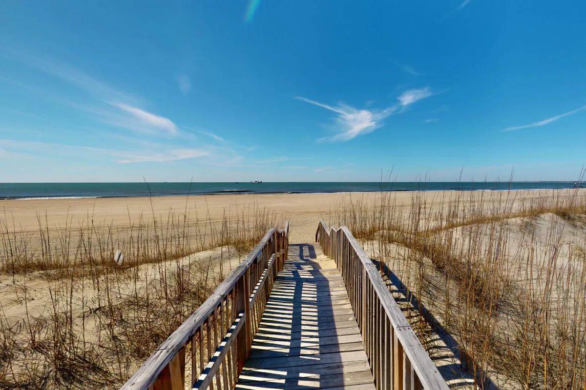 A wooden walkway leads out over the sand dunes toward a wide, inviting beach and the endless ocean horizon. Dune grass frames the path, and the soft lighting suggests early morning or late afternoon. The scene is tranquil, with no people visible, evoking a sense of private access and peaceful exploration.