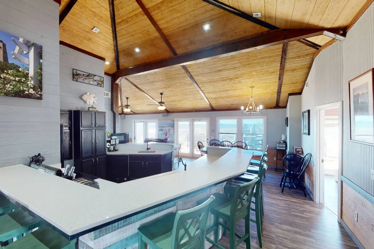 An inviting kitchen and bar area with a counter for casual seating, large windows letting in ample natural light, and distinctive wooden ceiling beams. The setup encourages both socializing and relaxed snacks or breakfasts, aligning with the central kitchen’s role in the home.