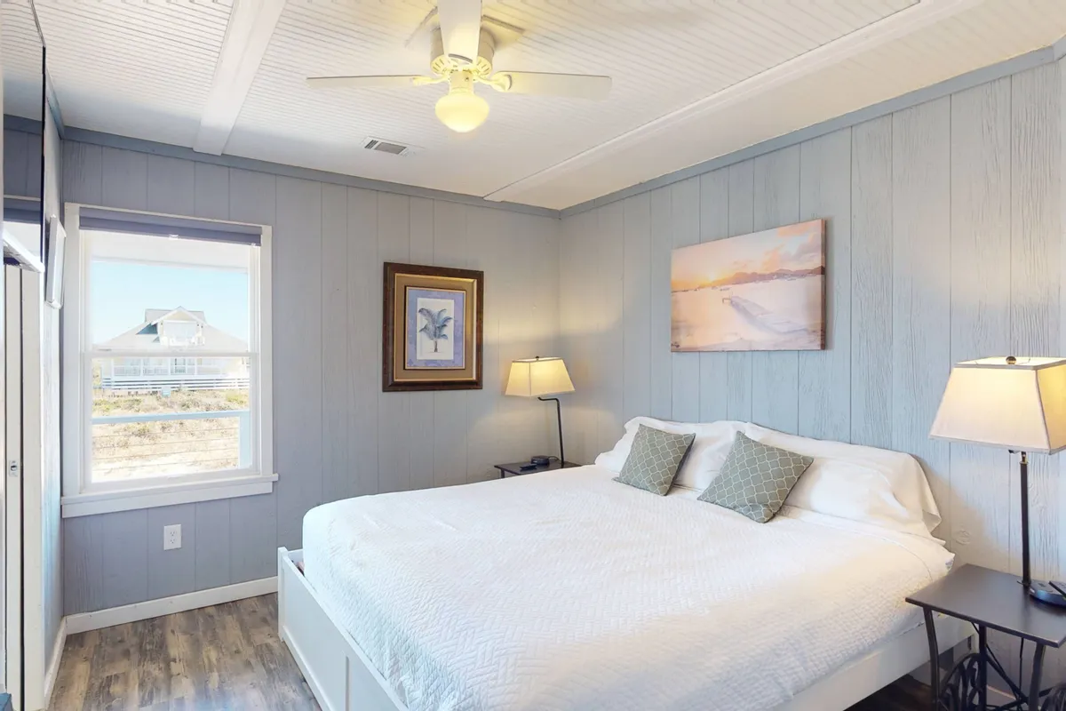 A serene king suite featuring a bed with crisp white linens, framed artwork, bedside table lamps, and a window offering soft natural light and a peaceful view.