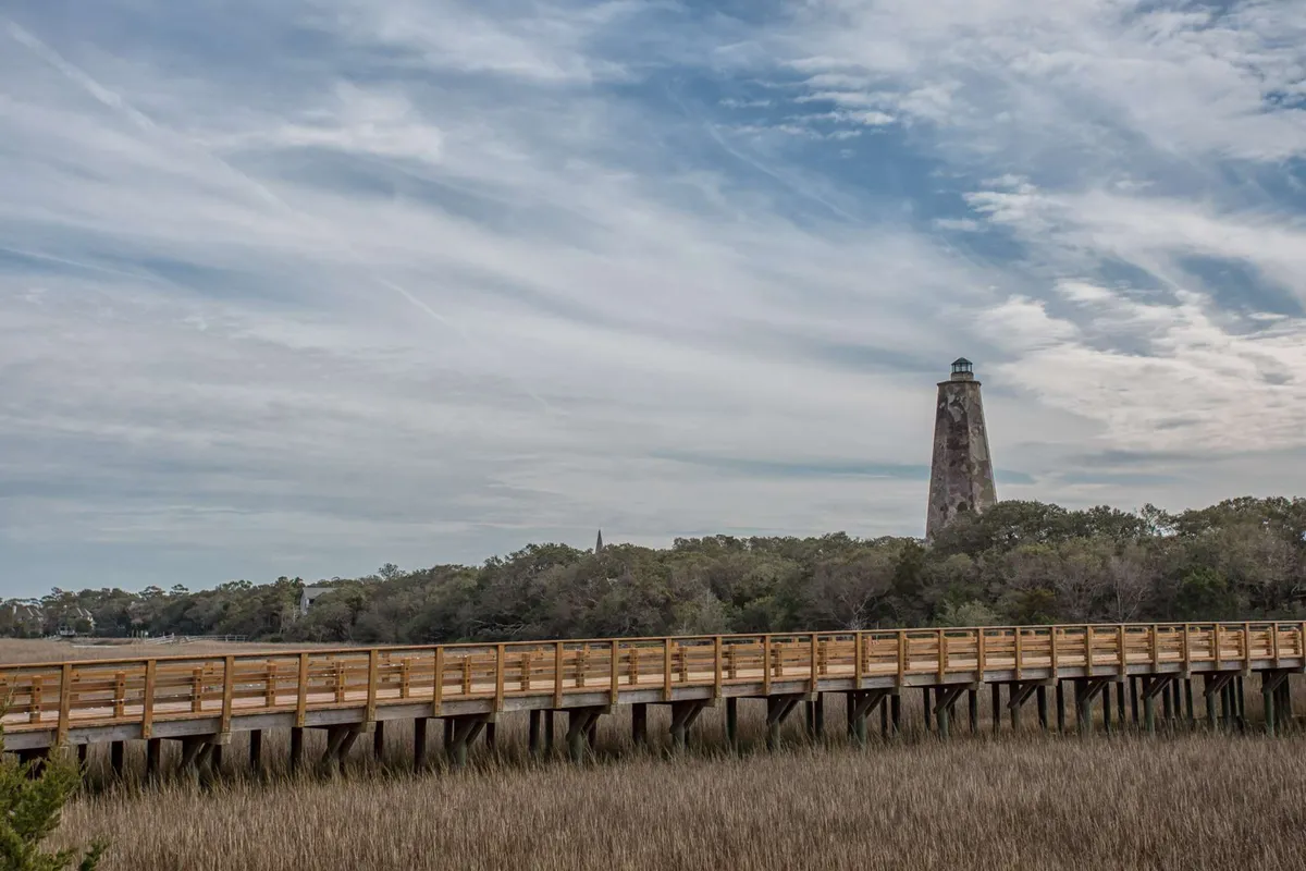 A wooden boardwalk winds through marshland toward a classic lighthouse, flanked by native grasses and open sky. The tranquil scene invites exploration, birdwatching, and photography—activities central to the section.