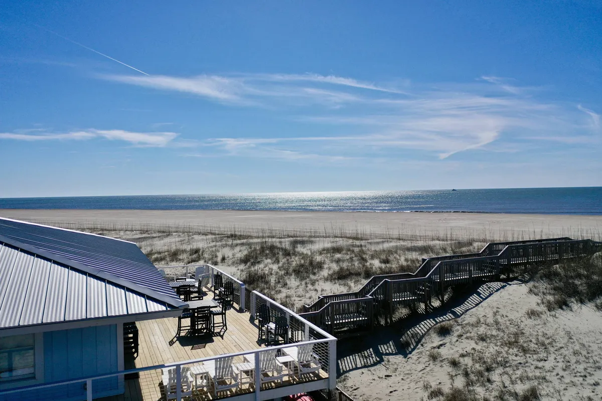 The image showcases a classic beach cottage perched atop the dunes, with a sprawling deck overlooking endless ocean views. The foreground features soft dune grass leading to a pristine sandy beach, bordered by the turquoise Atlantic. The deck is expansive, perfectly positioned for watching sunsets, enjoying sea breezes, and relaxing with friends and family. The scene is peaceful and inviting, with the ocean stretching out under a wide, open sky.