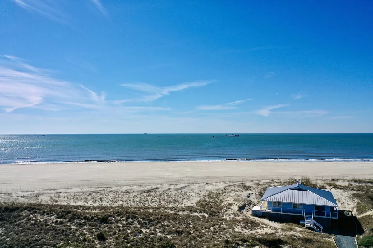A serene scene featuring a sunlit beach stretching alongside the ocean, with the beach house visible in the background. The environment looks peaceful and welcoming, perfect for long walks, play, or simply lounging together after a day of exploration.